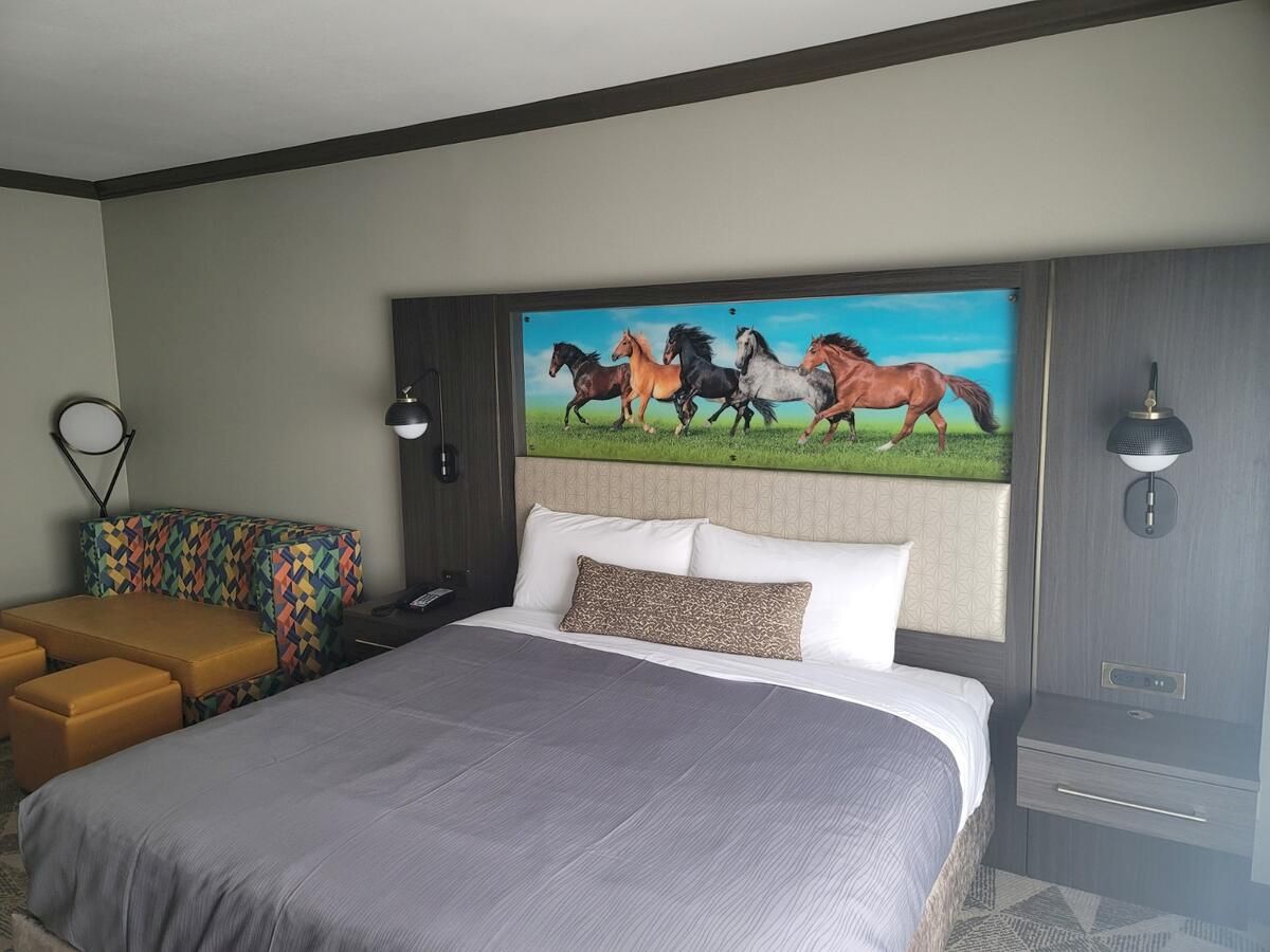A hotel room with a large bed, colorful couch, yellow ottoman, and a painting of running horses above the bed is shown.