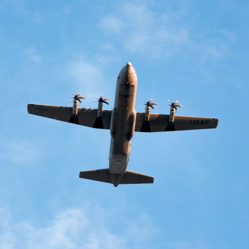 The image shows an aerial view of a four-engine propeller plane flying against a blue sky with some clouds.