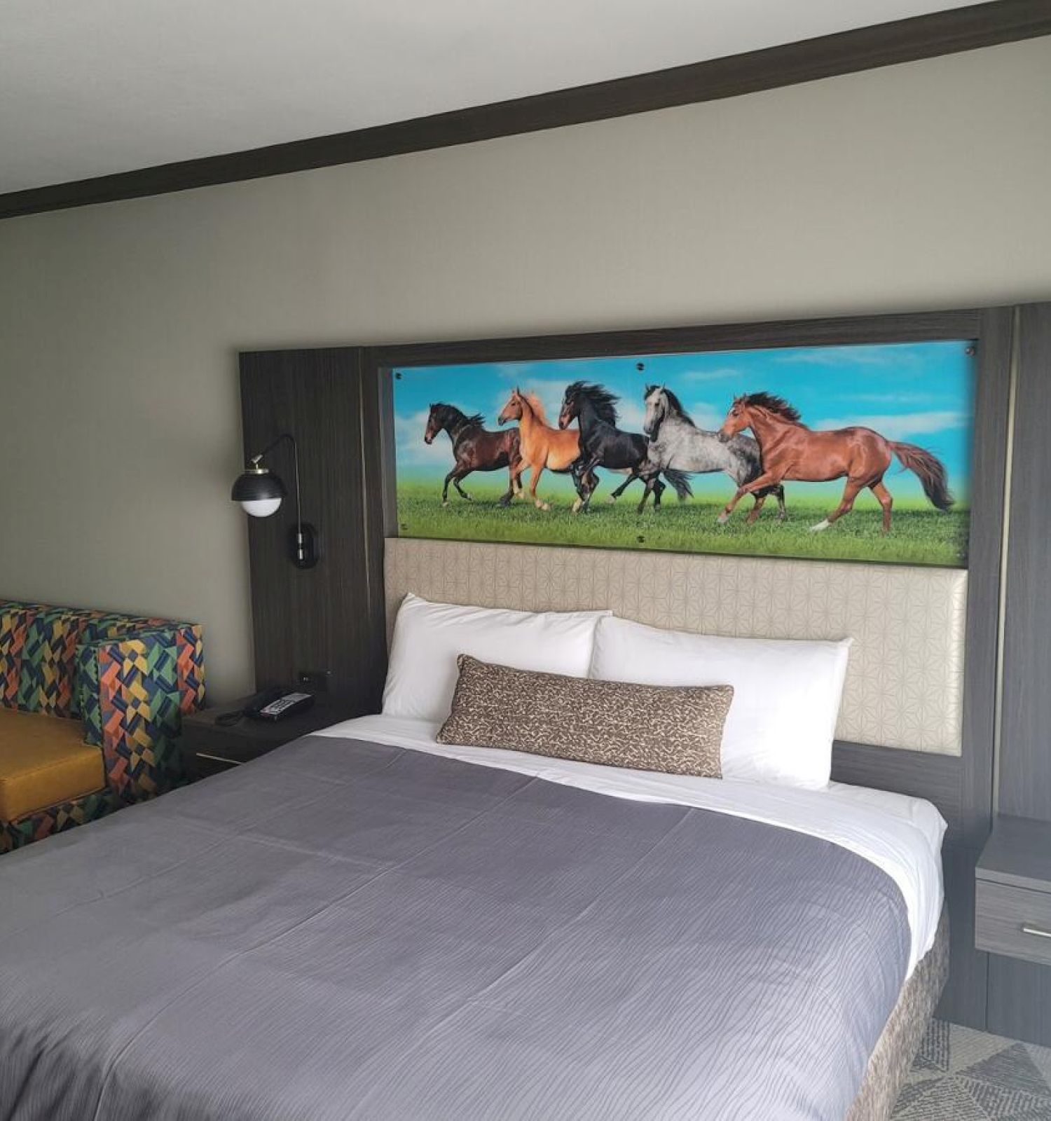 A hotel room with a large bed, a sofa, and a decorative painting of galloping horses above the bed. Two bedside lamps are present.