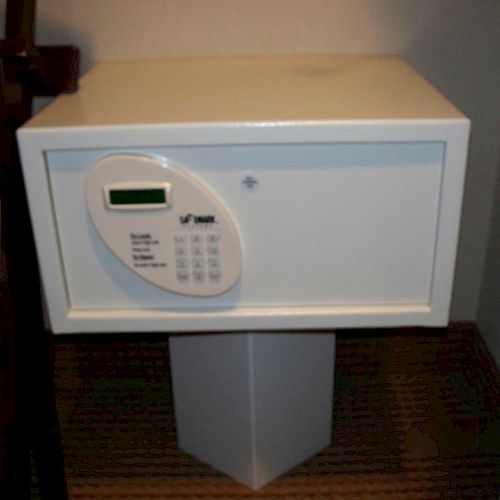 This image shows a white electronic safe with a keypad and a small display, placed on a pedestal in a closet or enclosed area.