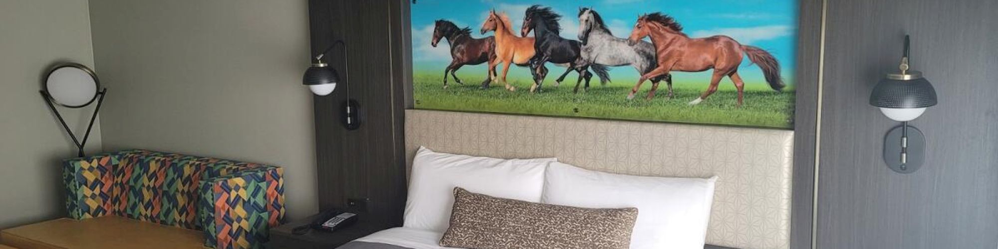A hotel room with a large bed, wall artwork featuring horses, a colorful sofa, two orange ottomans, bedside lamps, and a modern decor theme.