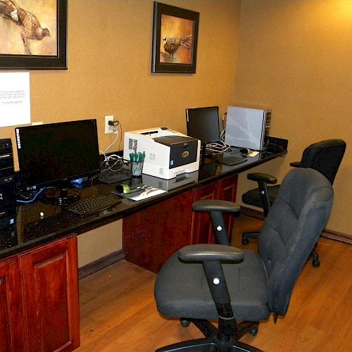 The image shows an office area with multiple computer stations, chairs, a printer, and artwork on the wall in a well-lit room, all arranged neatly.
