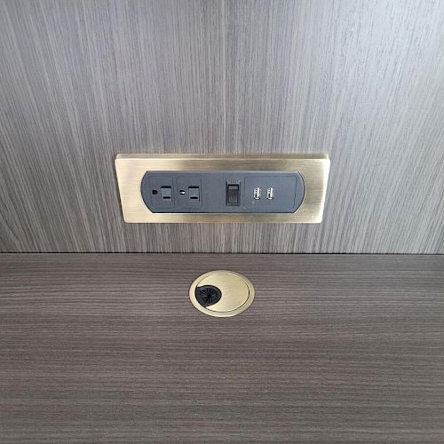 The image shows a power outlet with two electrical sockets and two USB ports installed on a wooden panel, with a cable management hole below it.
