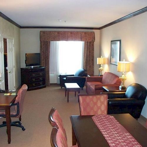 A hotel room features a big window, comfy chairs, lamps on side tables, a TV, a desk, and a dining table with chairs, creating a cozy atmosphere.