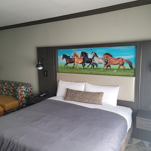 A neatly made bed with a gray comforter in a hotel room, wall art of running horses above the headboard, and a seating area on the left.