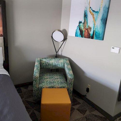The image shows a corner of a room with a green patterned chair, yellow ottoman, lamp, abstract wall art, and part of a bed with gray bedding.