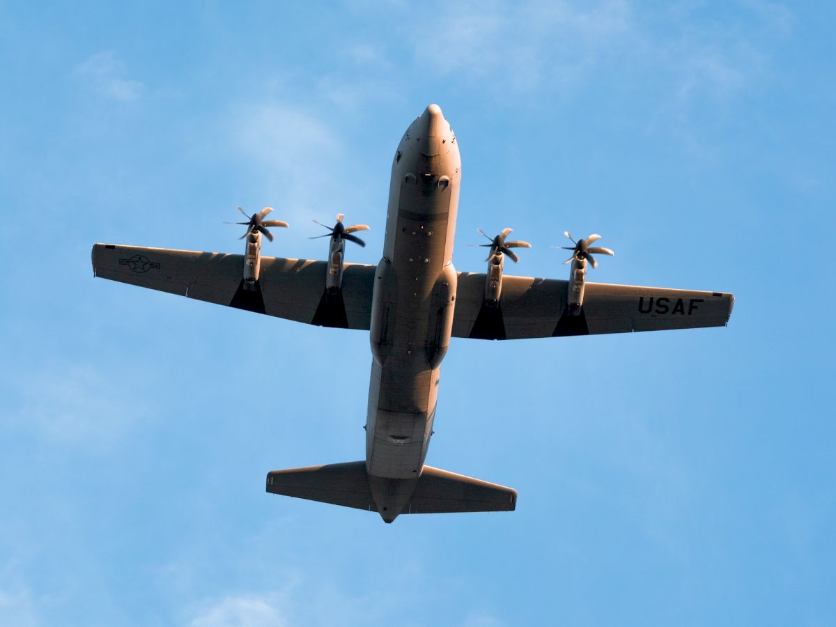 The image shows an aircraft with four propellers flying overhead against a blue sky with some scattered clouds.