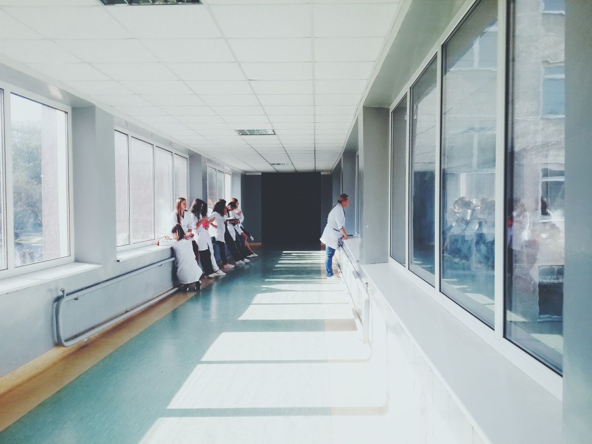The image shows a group of medical professionals in white coats standing in a bright, wide hallway with large windows in a hospital or medical facility.