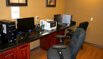 The image shows an office with computer workstations, office chairs, a printer, and framed artwork on the wall in a room with wooden flooring.