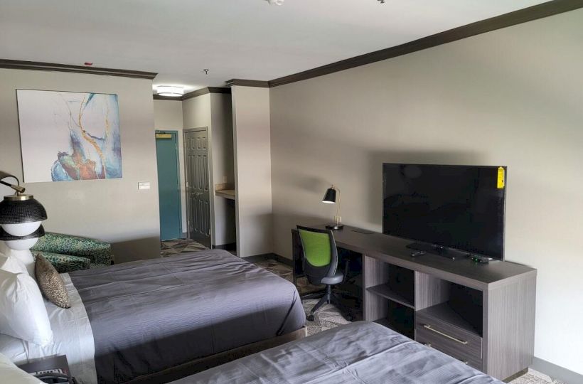 The image shows a hotel room with two beds, a TV on a stand, a chair, a desk lamp, an abstract painting on the wall, and a door in the background.