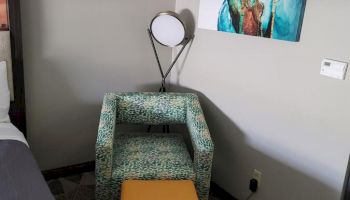 The image shows a chair with a yellow ottoman, an abstract wall picture, and a round table lamp in a corner of a room with patterned flooring.