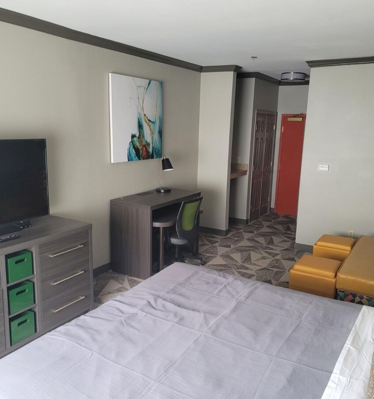 A modern hotel room features a bed, TV, dresser, desk with chair, colorful sofa, and abstract wall art. The room has neutral colors.
