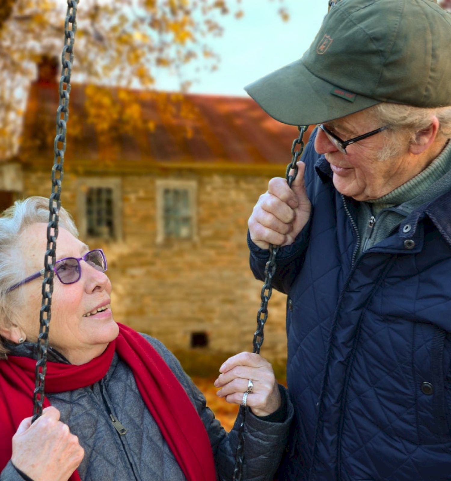 An elderly couple is standing outside in autumn, with the woman on a swing and the man holding its chains, smiling and looking at each other.