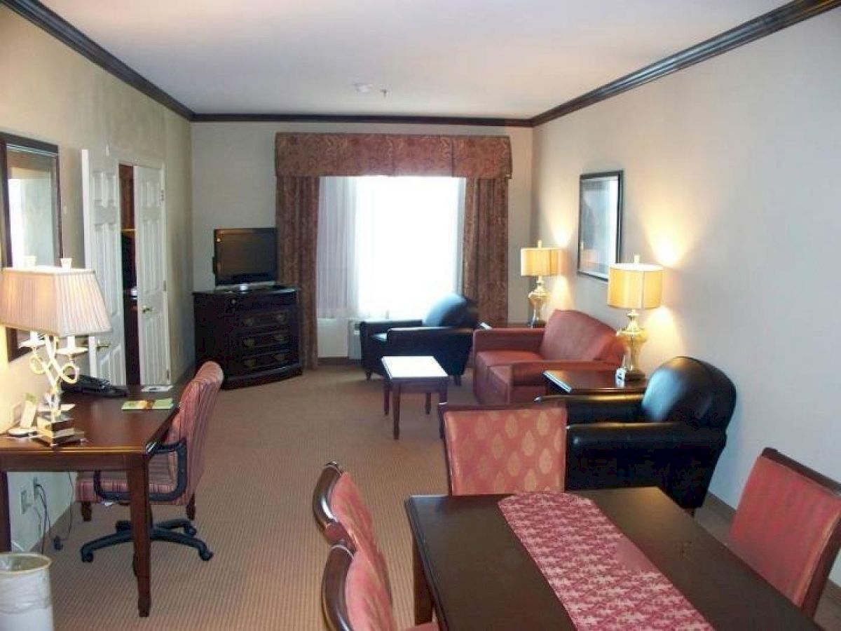 The image shows a spacious hotel room with a work desk, dining table, armchairs, a sofa, a TV, and lamps providing warm lighting.