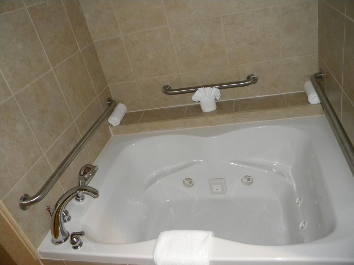 The image shows a clean, white bathtub with grab bars on the walls around it. There's a folded towel on the edge and a folded towel tucked.