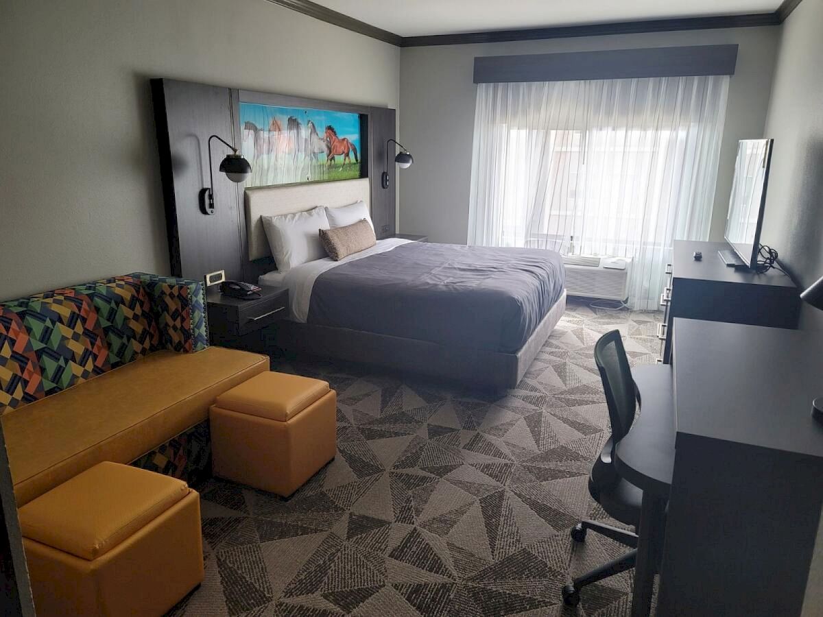 A modern hotel room features a bed, a small sofa with two ottomans, a desk with a chair, and a TV. The room has contemporary decor and lighting.