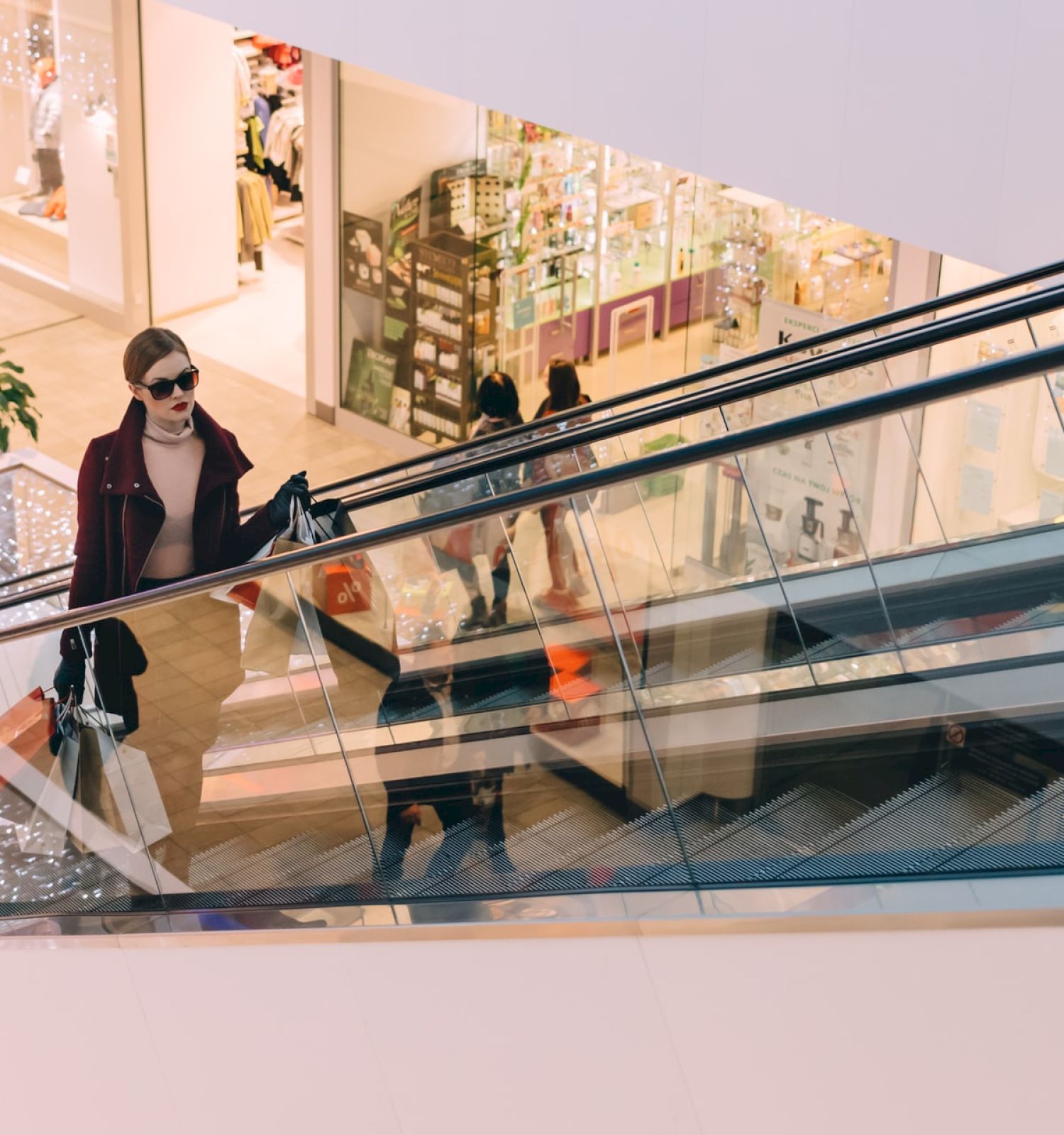 A person with sunglasses and a coat is on an escalator in a shopping mall, carrying shopping bags. The background shows various store fronts.