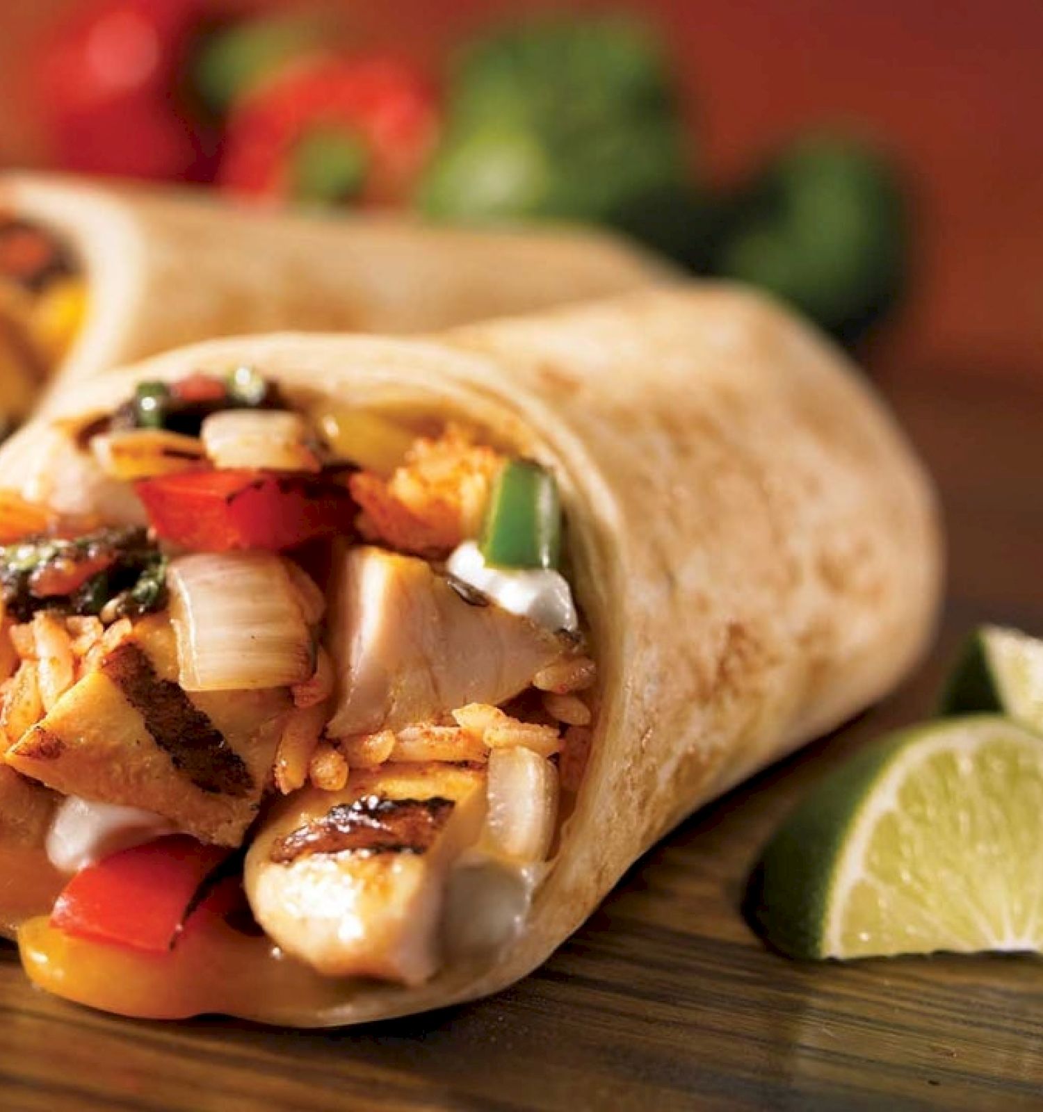 The image shows two burritos filled with vegetables, rice, and grilled chicken, accompanied by lime wedges, with a blurred background of tomatoes and peppers.
