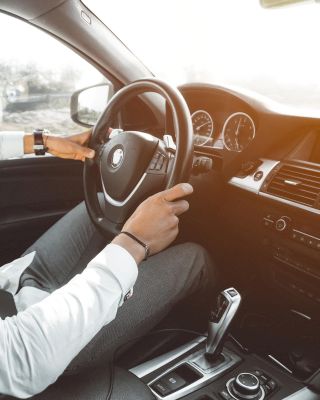 A person is driving a car, holding the steering wheel with their left hand and adjusting controls with the right, while sunlight streams through the window.