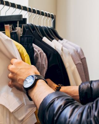 A person wearing a watch browses through a rack of clothes, holding a white t-shirt with 